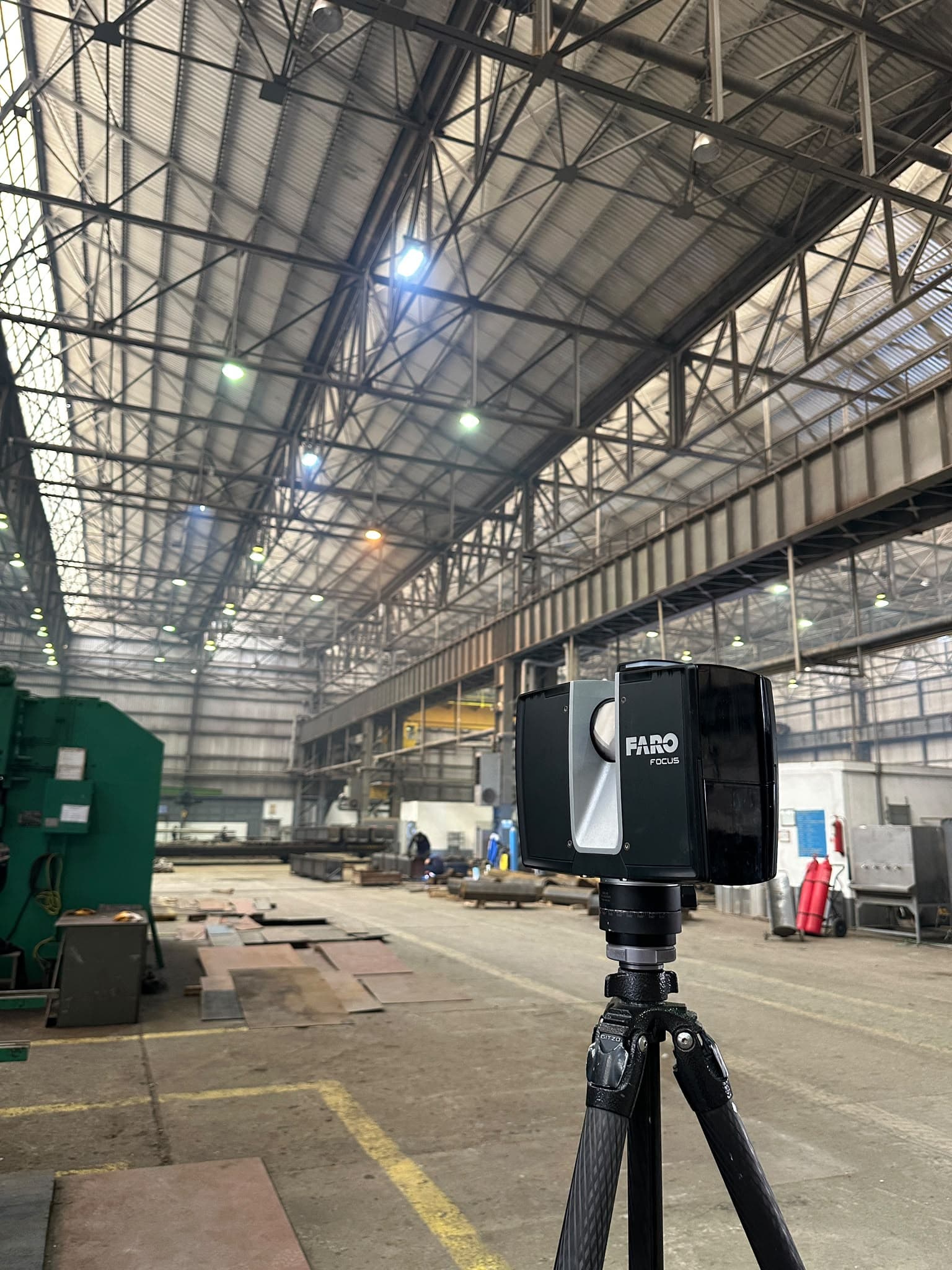 A FARO Focus laser scanner on a tripod inside an operating steel fabrication plant, with structural roof trusses and an overhead crane rail visible